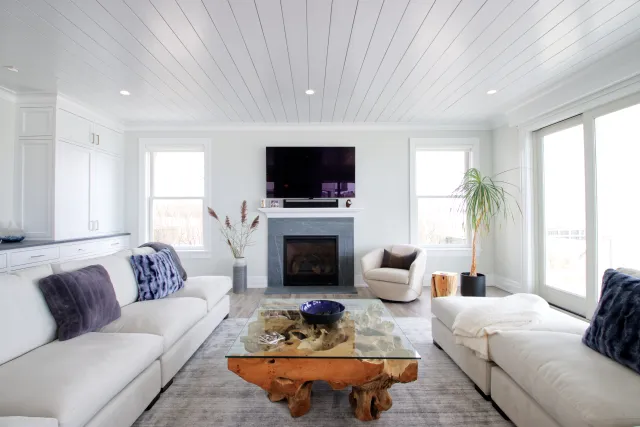 Bright modern living room with white sofas, glass coffee table, fireplace, and large windows letting in natural light