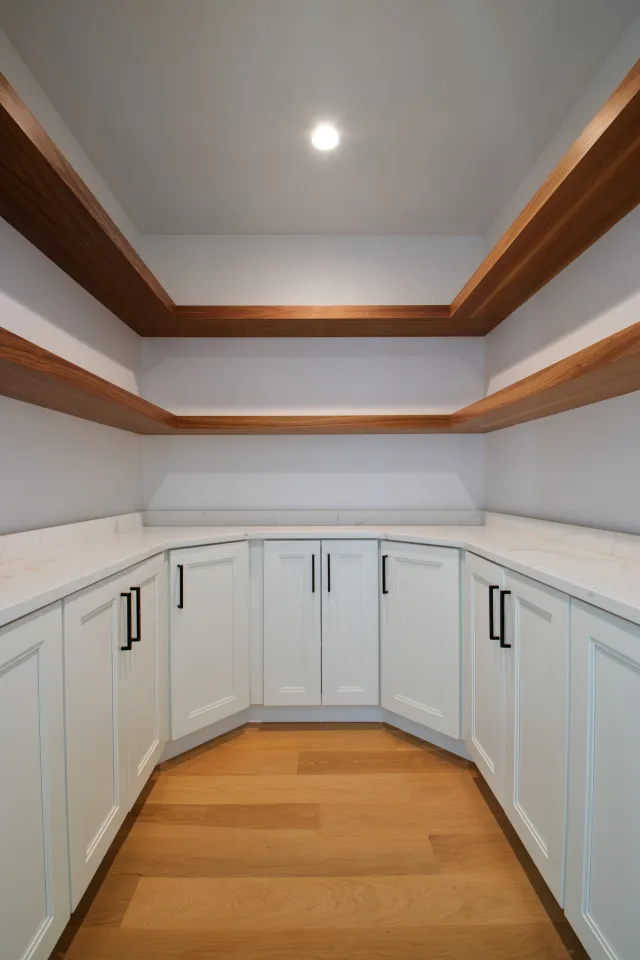Modern kitchen pantry with white cabinets, marble countertops, wood floating shelves, and hardwood floor.