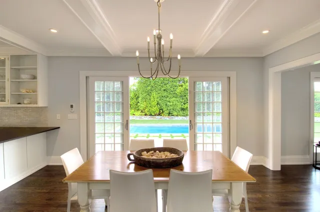 Bright dining room with wooden table, white chairs, chandelier, and glass doors opening to a pool and garden.
