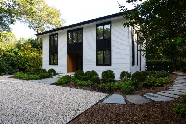 Modern white house with large windows, black accents, gravel driveway, and landscaped garden with stone path.