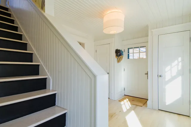 Bright entryway with white walls, black stair treads, wooden floors, and sunlight streaming through doors.