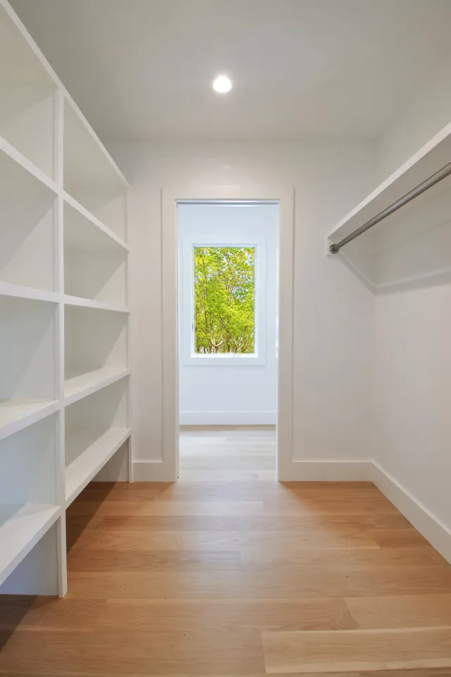 Bright walk-in closet with wooden floor, white shelves, hanging rod, and window showing green trees outside.