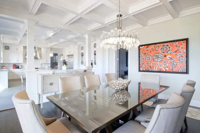 Modern dining room with glass table, beige chairs, chandelier, and colorful orange floral wall art.