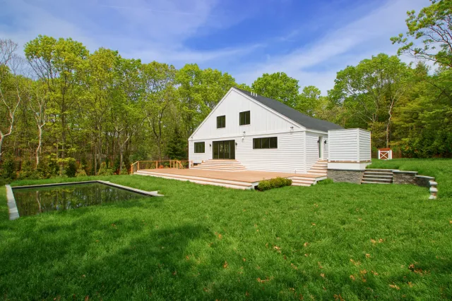 Modern white house with large wooden deck and small rectangular pond surrounded by green grass and trees.