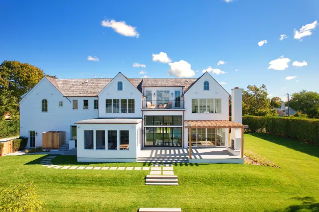 Large modern white house with multiple gable roofs, large windows, and spacious green lawn under a blue sky.