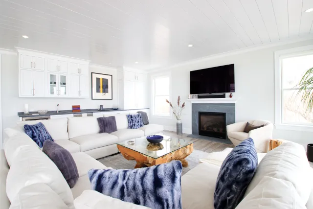 Bright living room with white sectional sofa, blue pillows, glass coffee table, fireplace, and wall-mounted TV