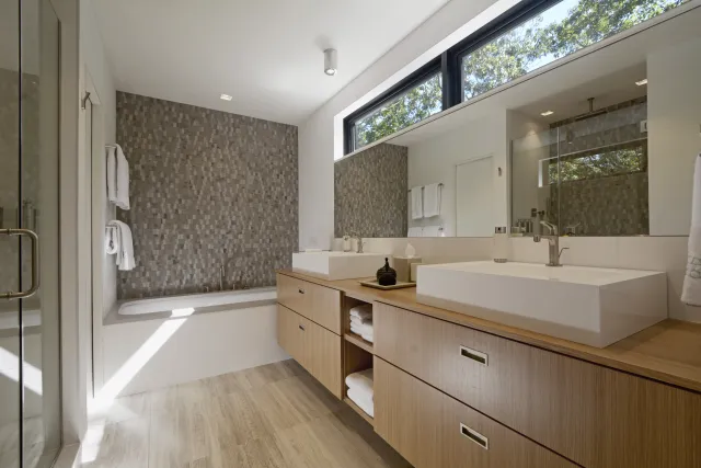 Modern bathroom with wood vanity, dual sinks, large mirror, textured tile wall, and natural light from high window.