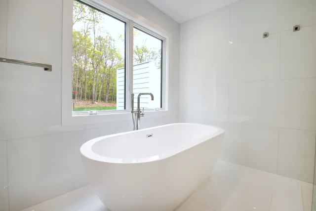 Modern white freestanding bathtub near large window overlooking trees in minimalist bathroom