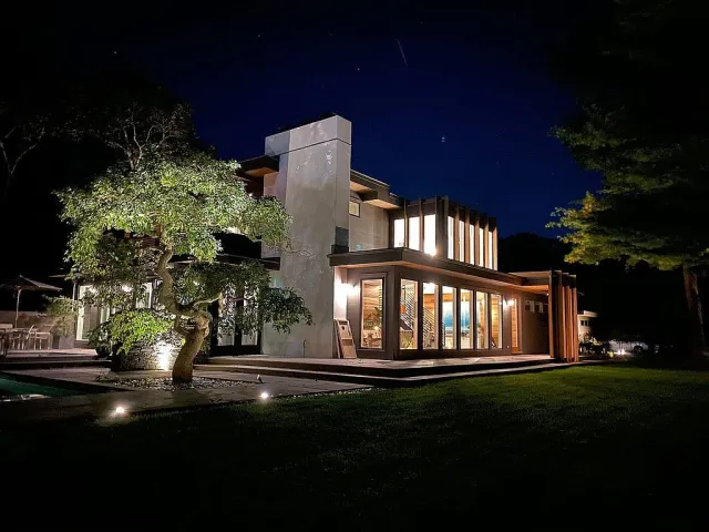 Modern house illuminated at night with large windows and a geometrically shaped tree in the yard.