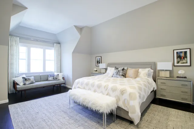 Bright modern bedroom with gray walls, large bed with plush pillows, furry bench, and window seat with cushions.