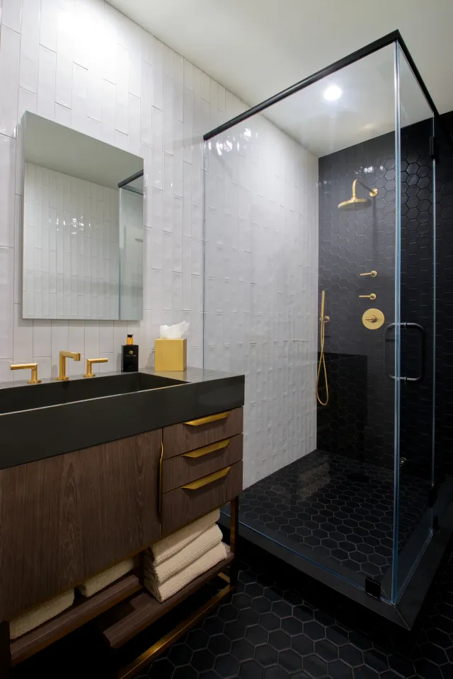 Modern bathroom with black hexagonal tiles, glass shower enclosure, dark wood vanity, and gold fixtures.