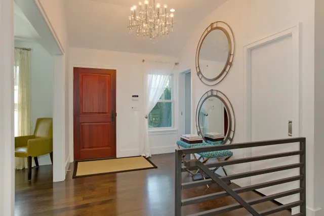 Bright entryway with wooden front door, two round mirrors, chandelier, and seating bench with books.
