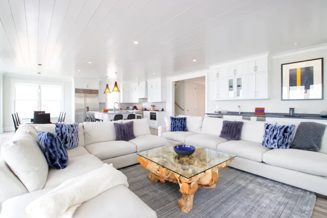 Bright open living room with white sectional sofa, glass coffee table, and modern kitchen in the background.