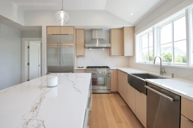Modern kitchen with stainless steel appliances, marble countertops, light wood cabinets, and large windows for natural light.