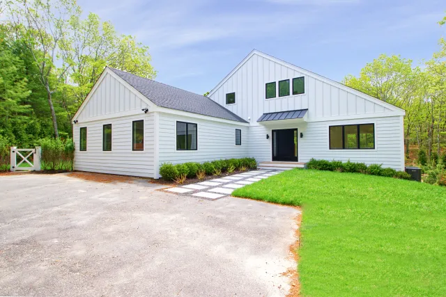 Modern white farmhouse with black windows, concrete driveway, green lawn, and surrounding trees under blue sky