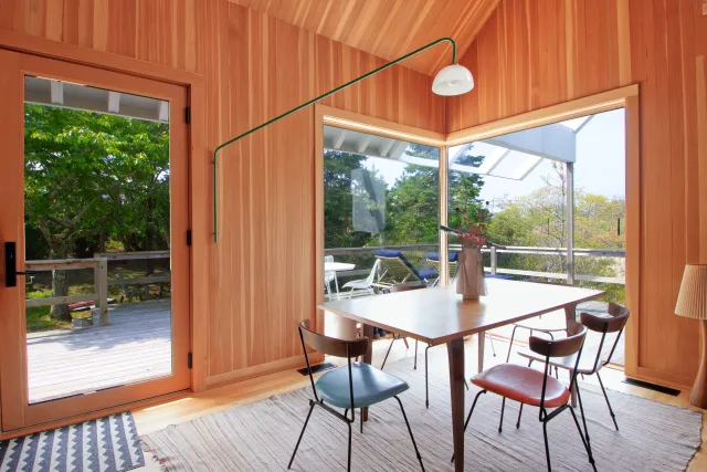 Sunlit dining area with wooden walls, table with mixed chairs, large glass doors opening to outdoor deck and greenery.
