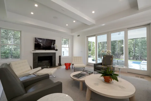 Bright modern living room with white walls, large windows, leather and white chairs, fireplace, and coffee tables.