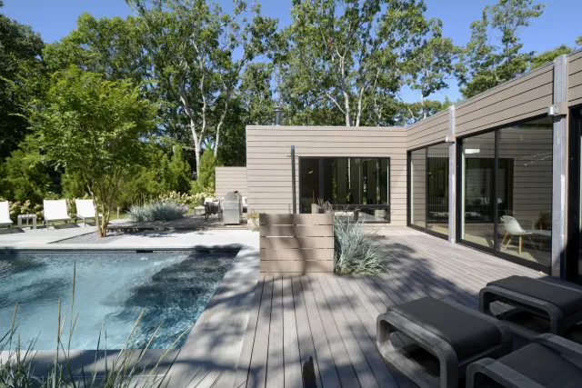Modern home patio with wooden deck, large glass windows, pool, lounge chairs, and surrounded by greenery on a sunny day