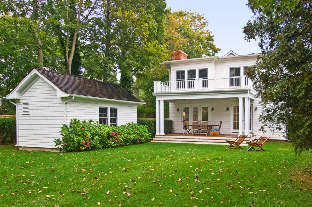 White two-story house with porch, balcony, green lawn, small shed, and surrounding trees in a suburban setting.