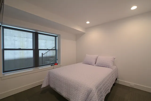 Minimalist bedroom with queen bed, light pink bedding, window with blind, and soft ceiling lighting.