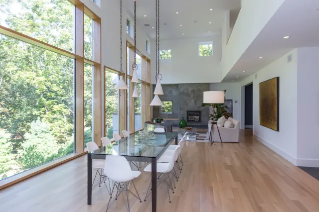Spacious modern dining area with large windows, glass table, white chairs, wooden floors, and a cozy living room.