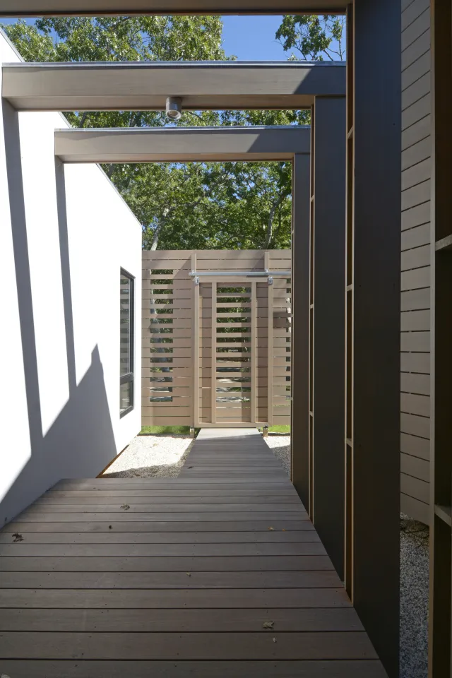 Modern outdoor wooden pathway leading to a horizontal slatted gate with white walls and green trees in background