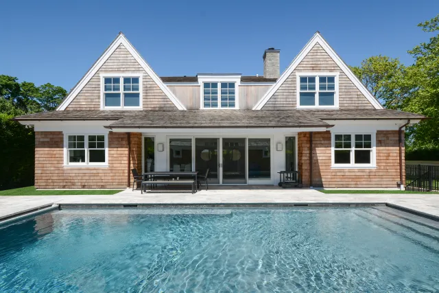 Modern wooden house with large windows and a swimming pool in front under a clear blue sky