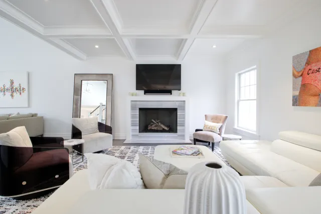 Bright modern living room with white furniture, fireplace, wall-mounted TV, and large window providing natural light