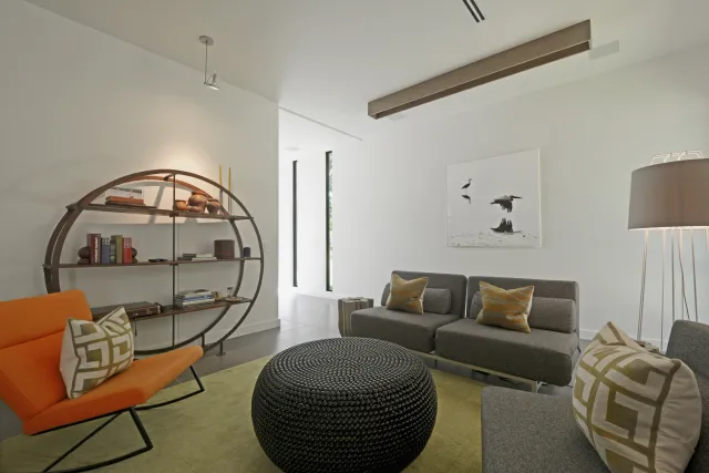 Modern living room with gray sofas, orange chair, round black ottoman, circular bookshelf, and floor lamp.
