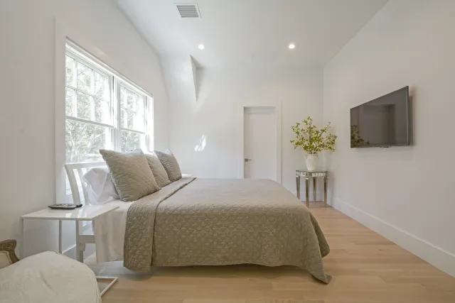 Minimalist bedroom with beige bedding, large window, wooden floor, wall-mounted TV, and a side table with plant.
