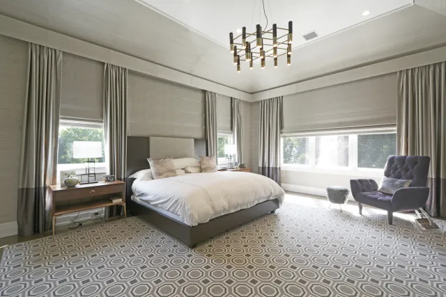 Spacious modern bedroom with large bed, geometric carpet, gray curtains, and a purple armchair under chandelier lighting.