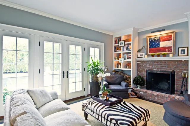 Bright living room with large French doors, brick fireplace, zebra print ottoman, gray sofa, and built-in bookshelves.