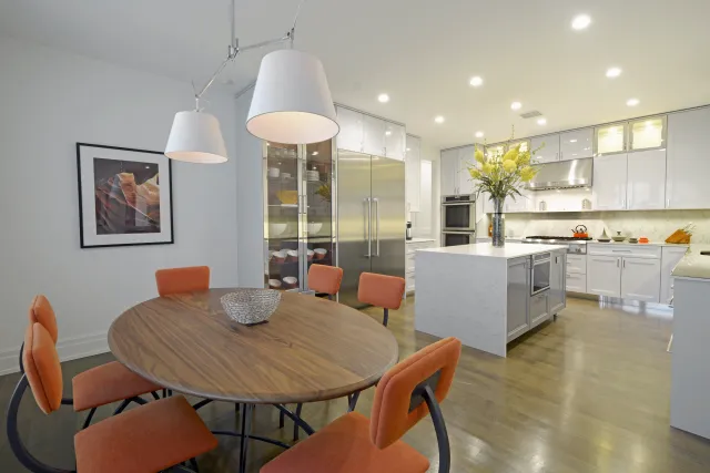 Modern kitchen and dining area with white cabinets, marble island, stainless steel appliances, and orange chairs.