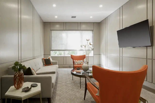 Modern living room with beige sofa, orange armchairs, round coffee table, wall-mounted TV, and large window.