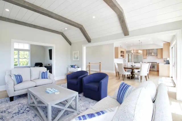 Bright living room with gray sofas, blue armchairs, wooden beams, open dining area, and light hardwood floors