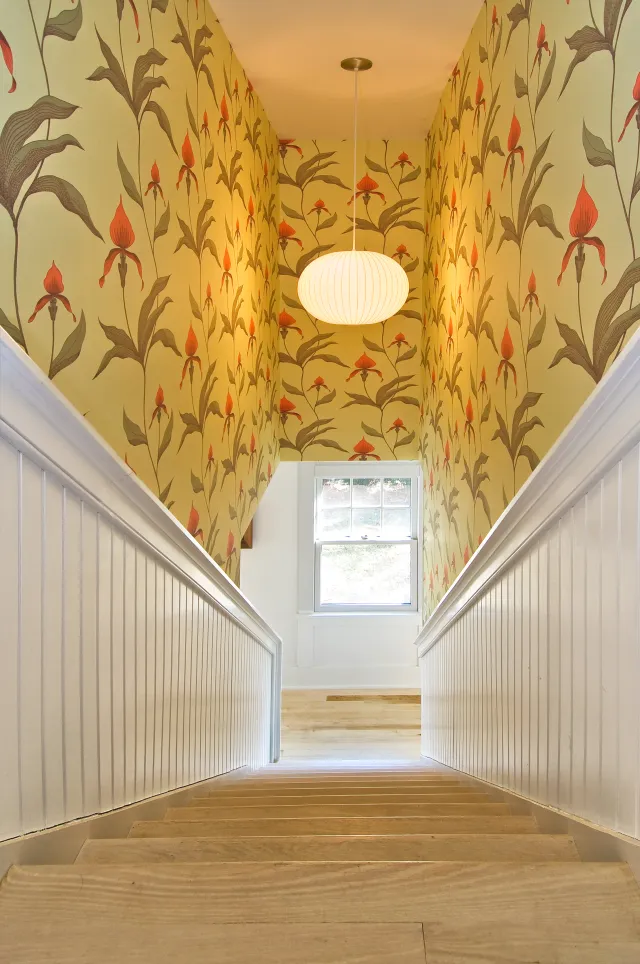 Staircase with wooden steps leading up to a window, floral wallpaper, white wainscoting, and a round ceiling light.
