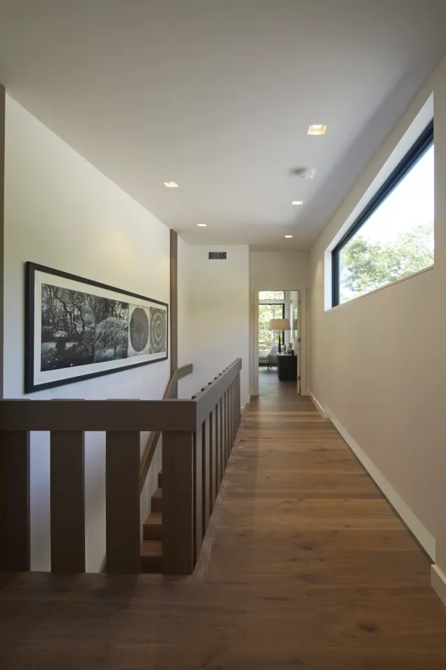 Modern hallway with wooden floor, large window, staircase railing, and framed black-and-white artwork on wall.