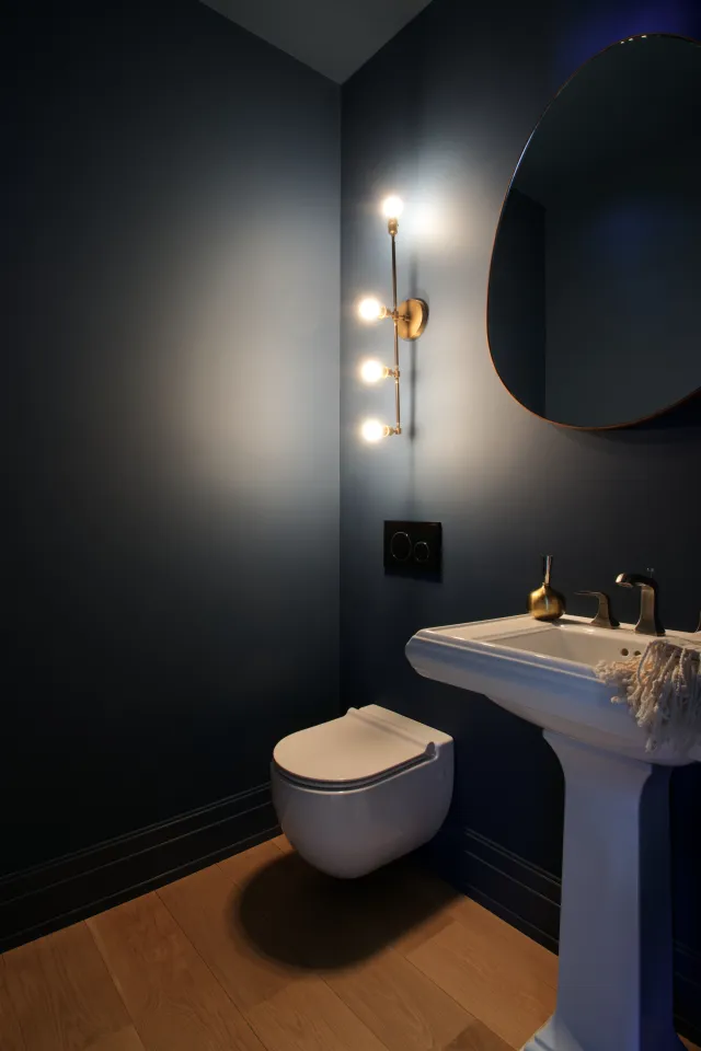 Modern bathroom with navy walls, wall-mounted toilet, pedestal sink, round mirror, and vertical light fixture.