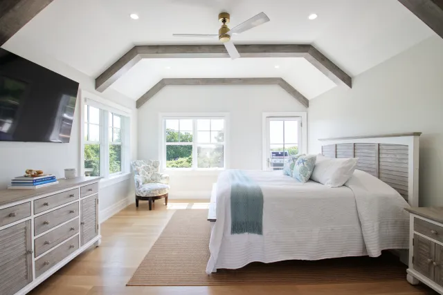 Bright white bedroom with wooden beams, queen bed, cozy armchair, ceiling fan, and large windows letting in natural light