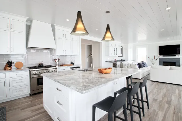 Bright modern kitchen with white cabinetry, marble island, black bar stools, and open living area with sofa and TV.
