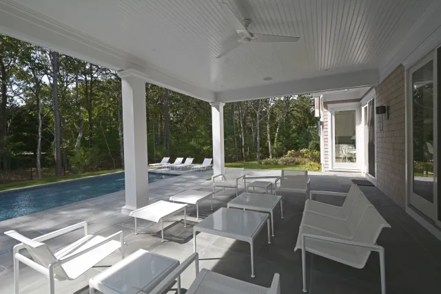 Modern outdoor patio with white chairs and tables overlooking a swimming pool and forested backyard.