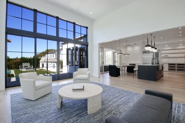 Modern living room with large windows, white armchairs, grey sofa, round coffee table, open kitchen in background