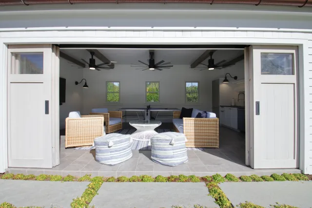 Modern outdoor lounge area with wicker chairs, poufs, ceiling fans, and a white table inside a white patio space.