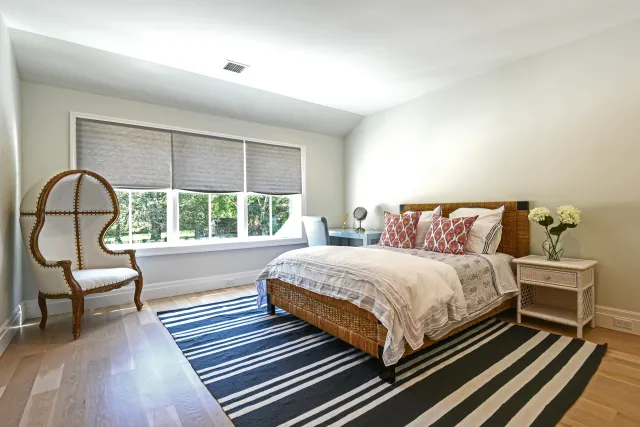 Bright bedroom with wicker bed, white linens, patterned pillows, striped rug, elegant chair, and large window with blinds