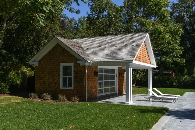 Small backyard pool house with shingle siding, white columns, and lounge chairs on tiled patio under a sunny sky.