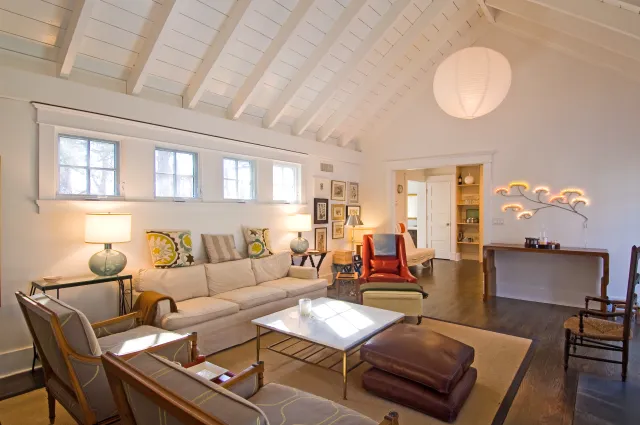 Bright living room with vaulted ceiling, cream sofa, patterned cushions, leather chair, and modern lighting fixtures