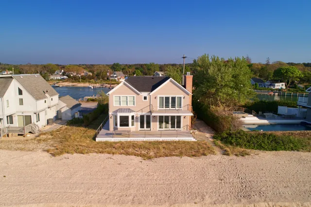 Beach house with large windows overlooking sandy shore and calm water under clear blue sky.