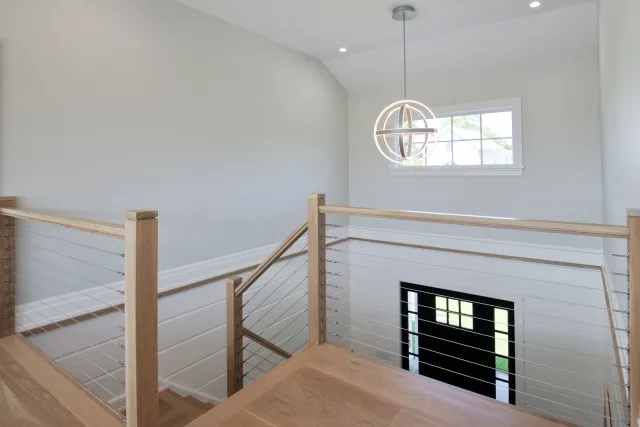 Modern staircase with wooden handrails, metal cable balusters, pendant light, and black front door with window.
