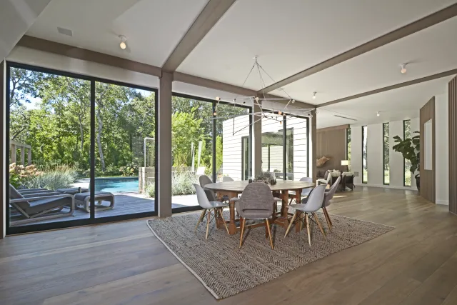 Modern dining area with round table, gray chairs, large windows overlooking a deck and swimming pool.