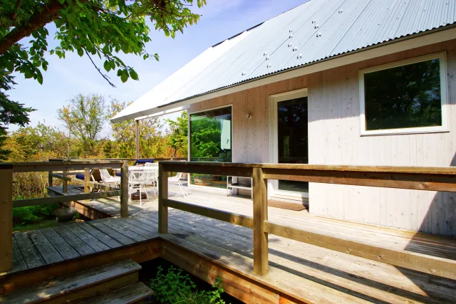 Wooden deck with railing attached to a modern white house surrounded by trees on a sunny day